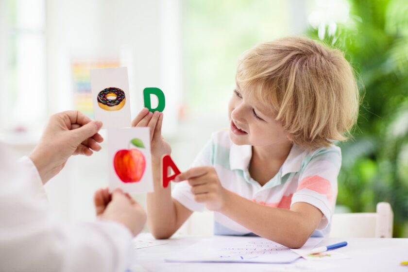 A child smiles while holding a letter "A" card with an apple image, sitting opposite an adult holding a "D" card with a donut image. Bright, airy room in the background.