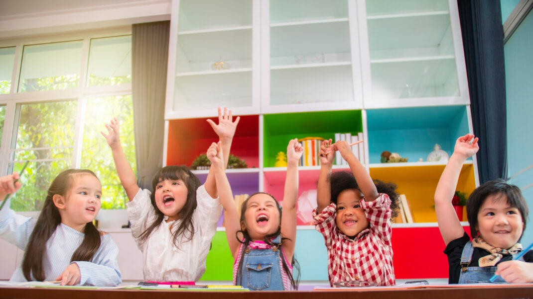 Five children seated at a table with colorful background shelves, all raising their hands enthusiastically, smiling, and appearing excited.