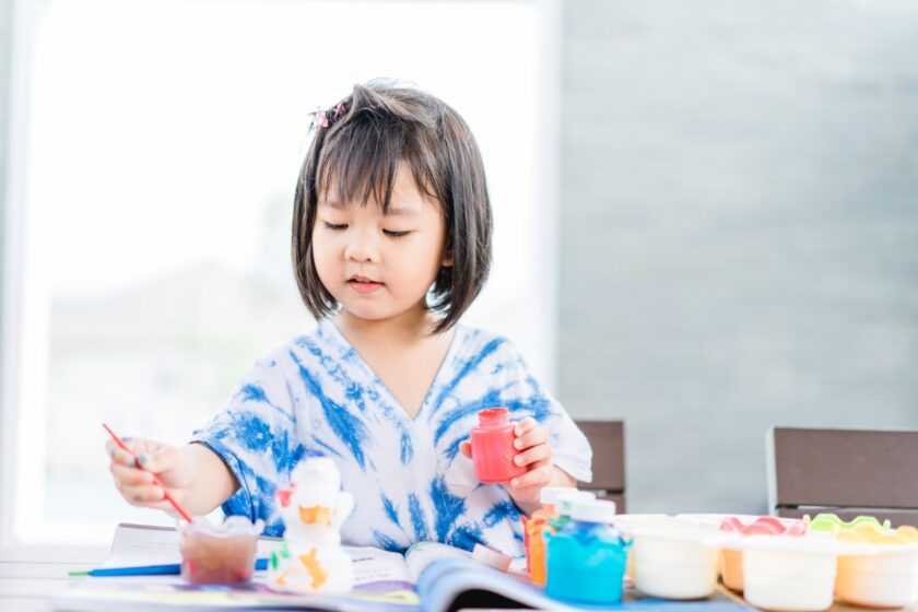 A young girl sitting at a table is painting with colorful paints, holding a red paint container in one hand and a brush in the other. Various paint pots are scattered on the table.