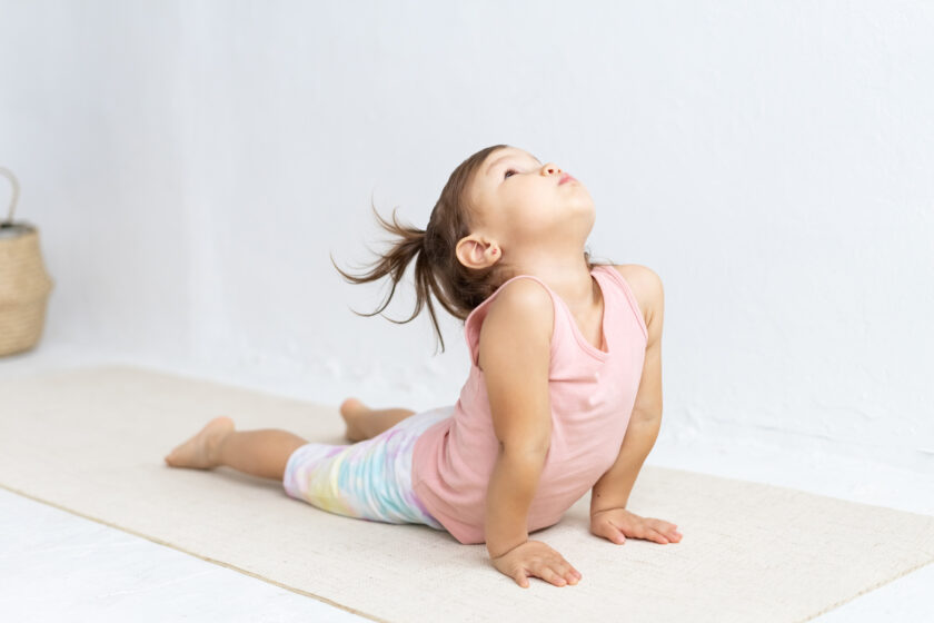 A young child with pigtails performs a yoga cobra pose on a mat, wearing a pink sleeveless top and colorful leggings against a white background.