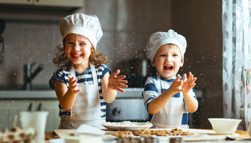 Two children wearing chef hats and aprons are joyfully clapping their hands, causing flour to fly in a kitchen. Various baking ingredients and utensils are spread out on the table in front of them.